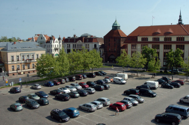 na zdjęciu widzimy plac, kilkadziesiąt samochodów zaparkowanych, kilka drzew, w tle jednię i inne budyni